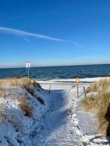 Weg durch Sand und Schnee zum Meer, blauer Himmel und ruhiges Wasser im Hintergrund.